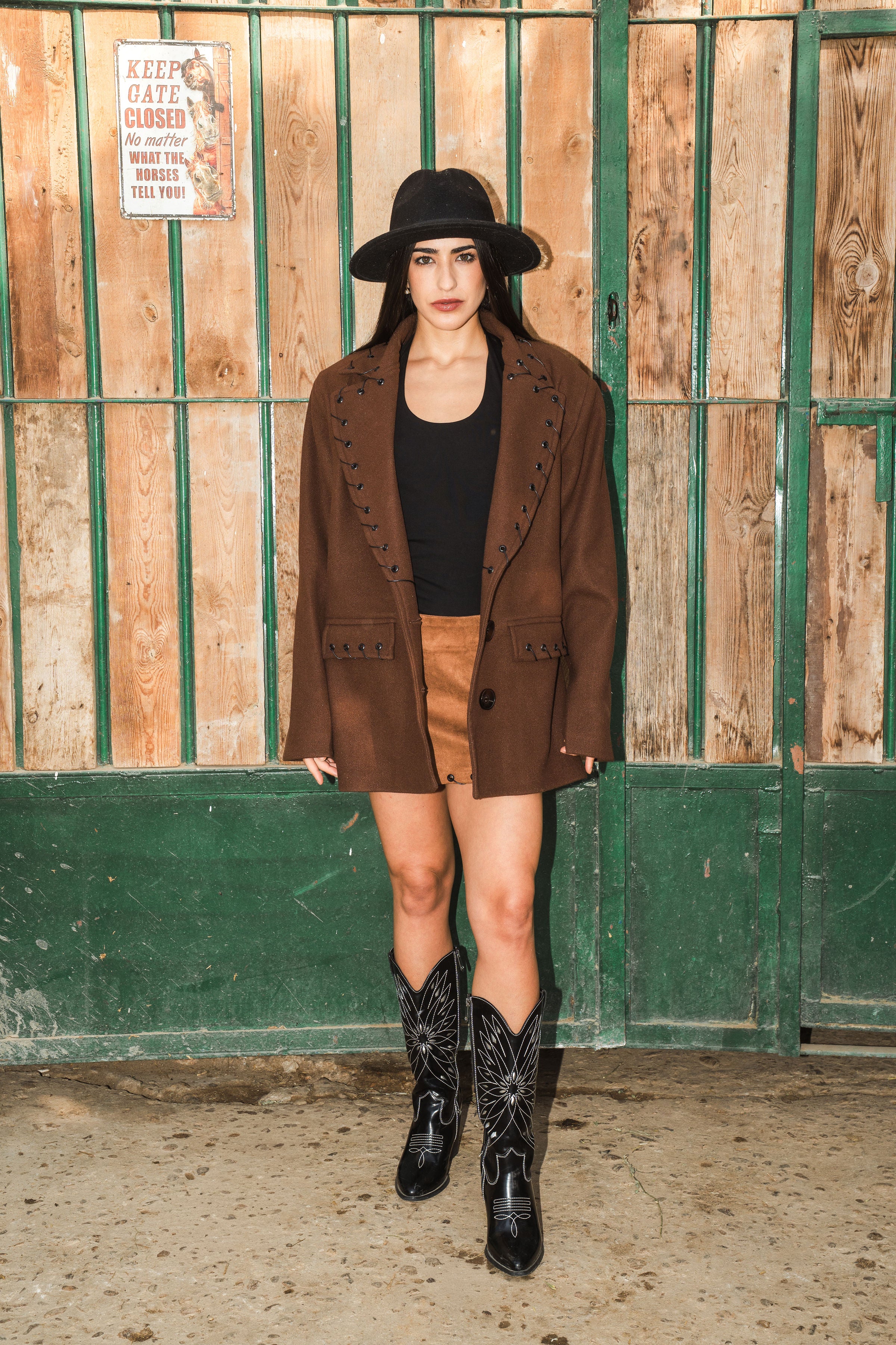 Woman in a brown coat, black top, and tan skirt standing in front of a wooden gate.