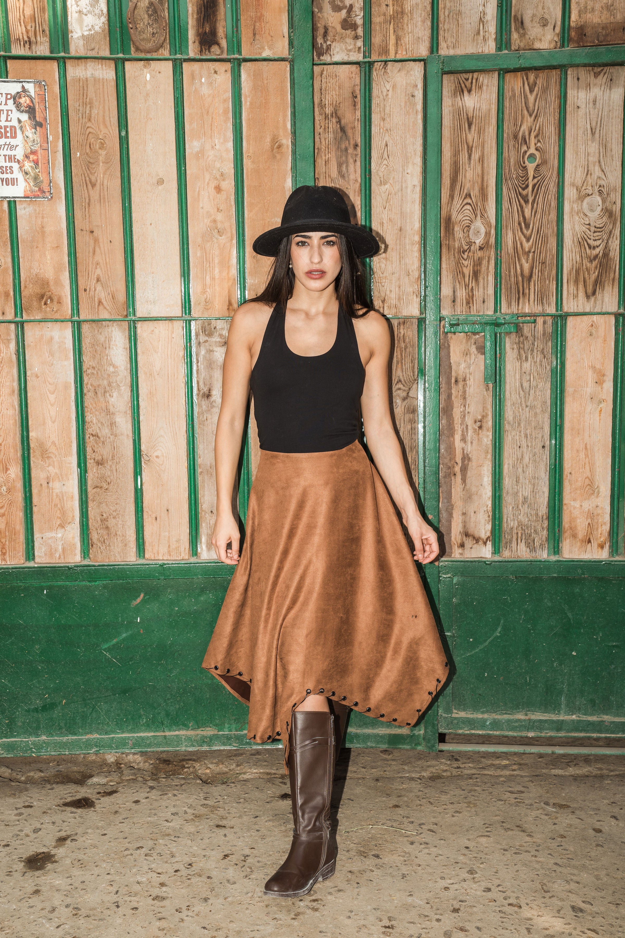 Woman wearing a black top, brown skirt, and brown boots standing against a wooden fence.